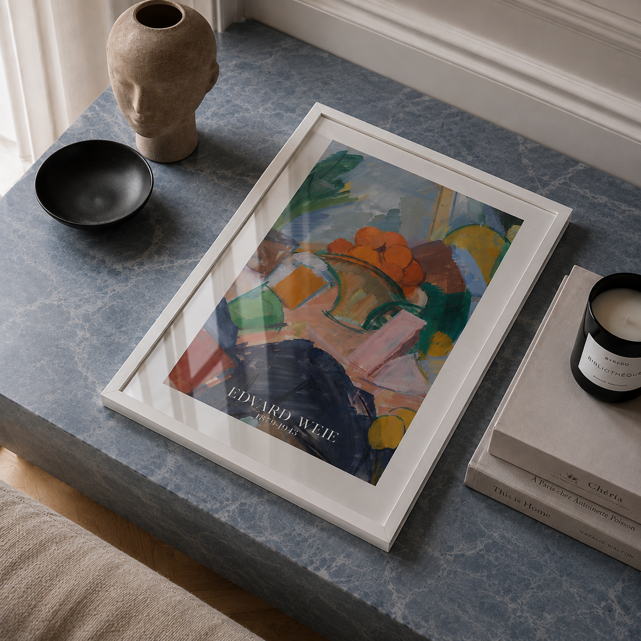 A framed art print featuring an abstract still life painting with the text 'EDVARD ALDEME' and 'BELGIUM' at the bottom, styled on a blue marble surface next to a black bowl, a ceramic head sculpture, and books.