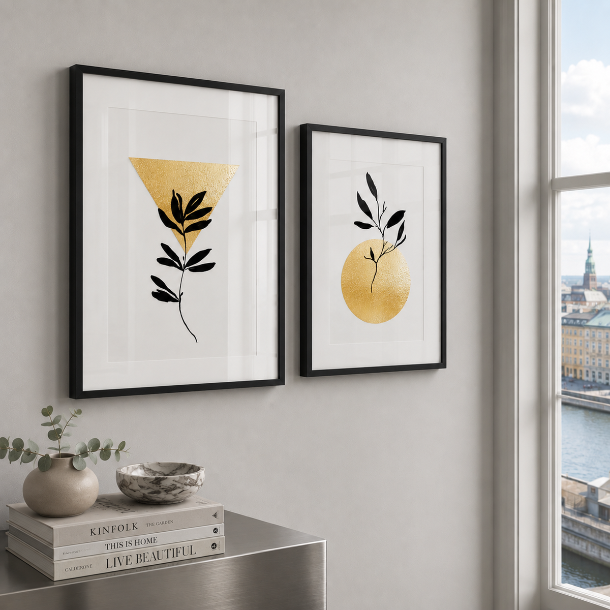 Two framed minimalist art prints with black frames hang on a light gray wall in a modern room. The left print features a black leaf motif inside a gold triangle on a white background, and the right print features a black leaf motif inside a gold circle.