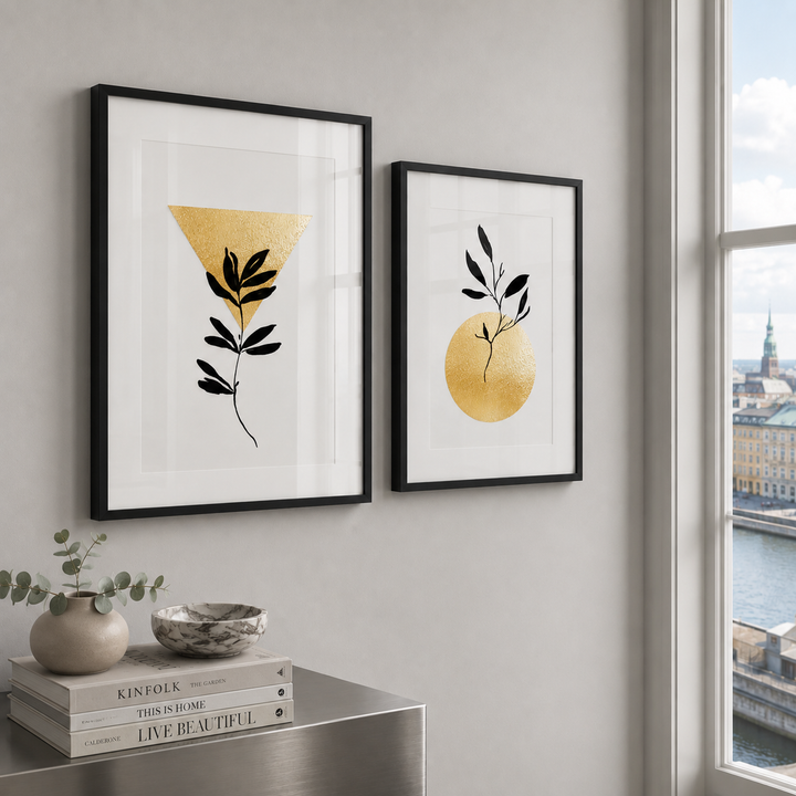Two framed minimalist art prints with black frames hang on a light gray wall in a modern room. The left print features a black leaf motif inside a gold triangle on a white background, and the right print features a black leaf motif inside a gold circle.