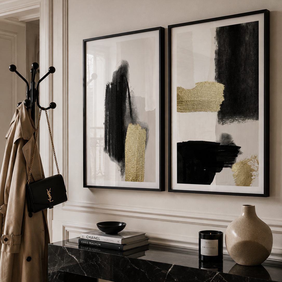 Two framed abstract wall art prints with black and textured gold brushstrokes on a white background, displayed in a styled interior hallway above a black marble console table.