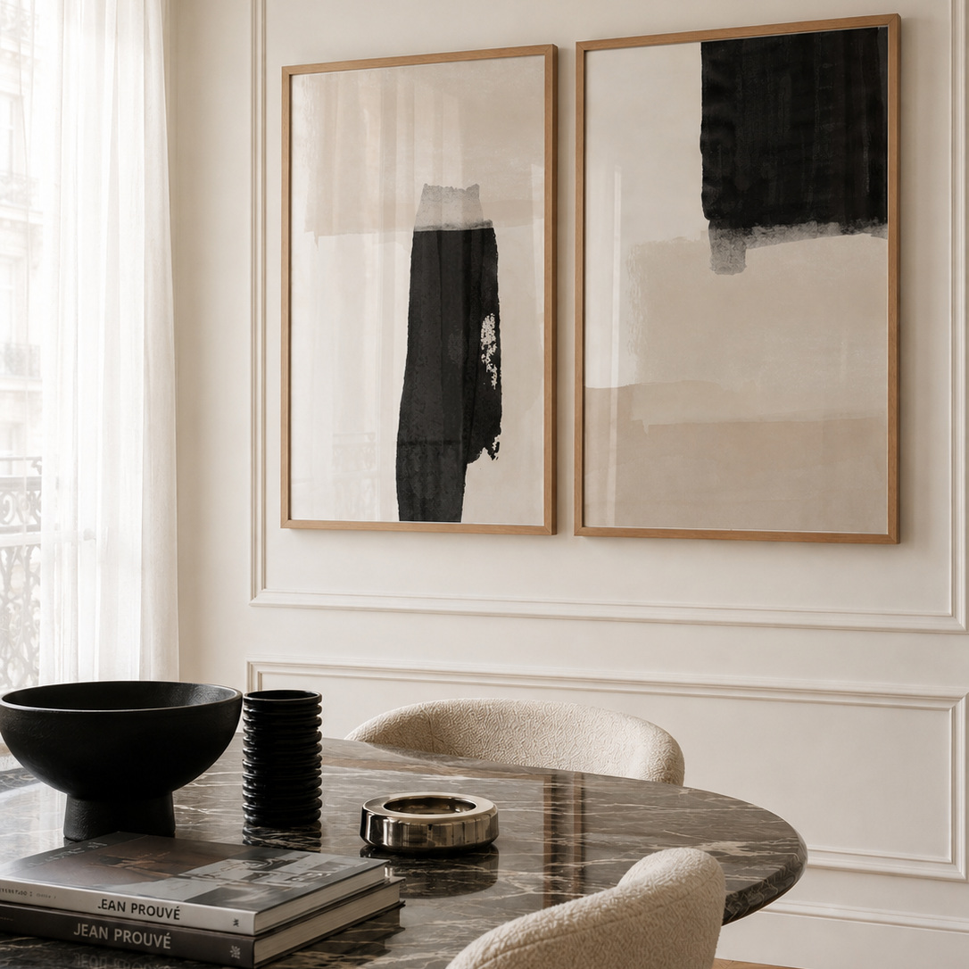 Two framed abstract black and beige canvas prints hang on a white paneled wall above a dark marble dining table with neutral upholstered chairs.