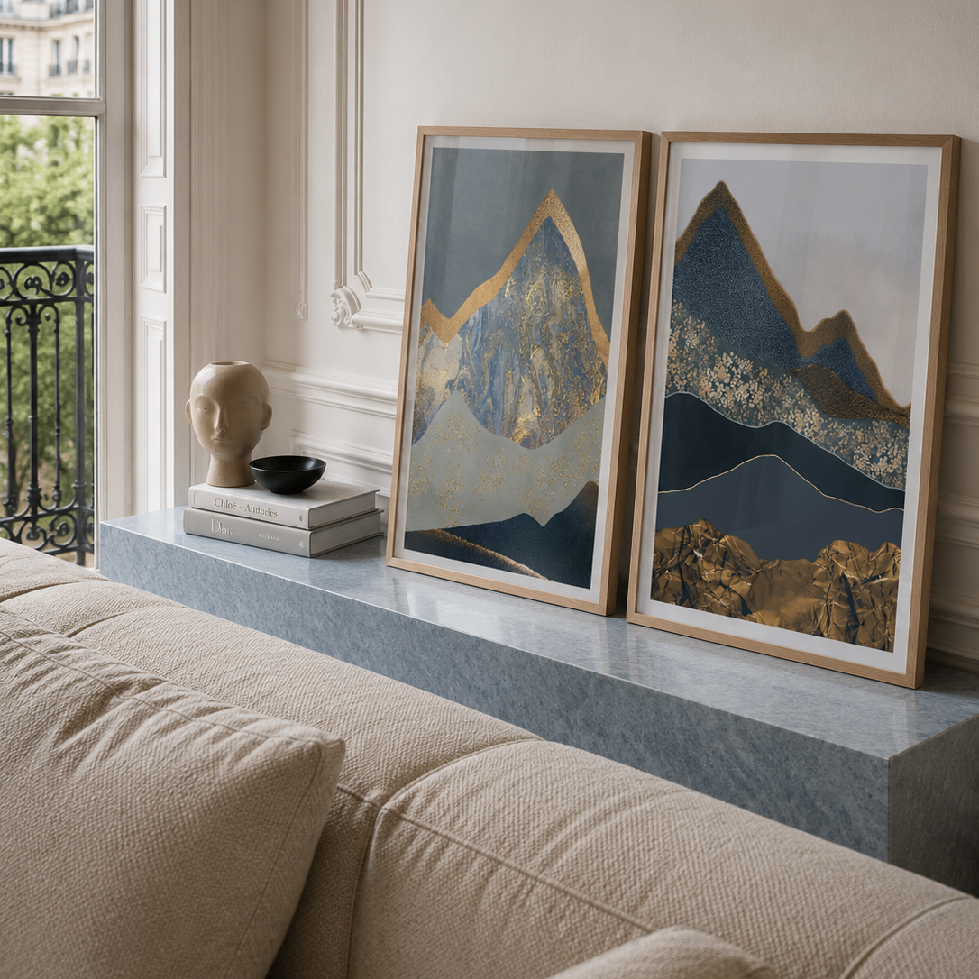 Two framed abstract mountain art prints with gold, navy, and gray tones displayed on a gray marble mantelpiece above a beige sofa in a styled living room.