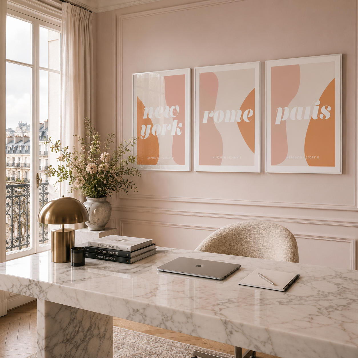 Three framed abstract art prints with city names 'new york', 'rome', and 'paris' hang on a blush pink wall above a marble desk in a bright, elegant room.