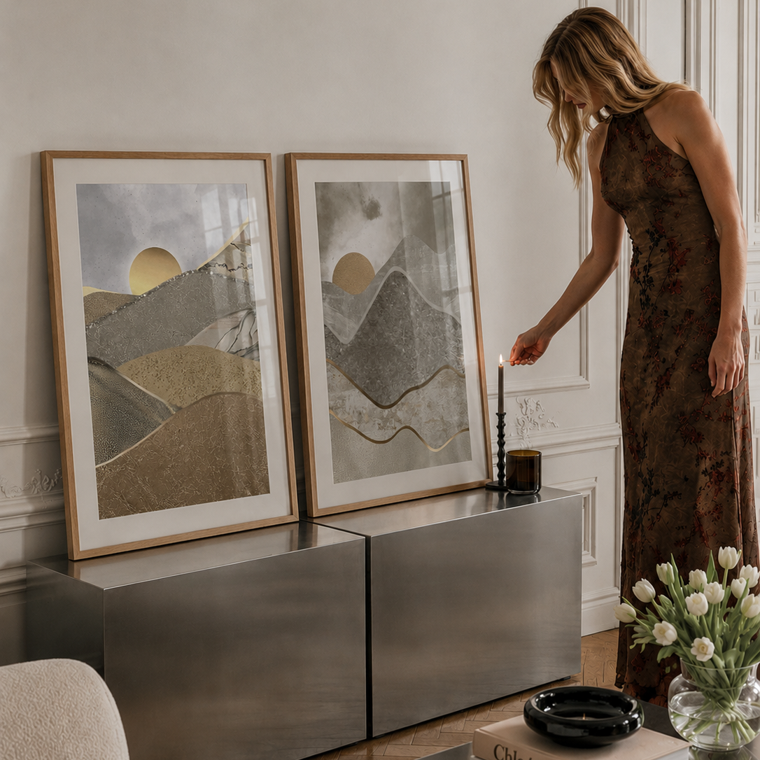 A woman in a brown floral dress stands beside a metallic console table in a styled living room, lighting a candle next to two large framed abstract landscape prints with gold, gray, and beige tones.