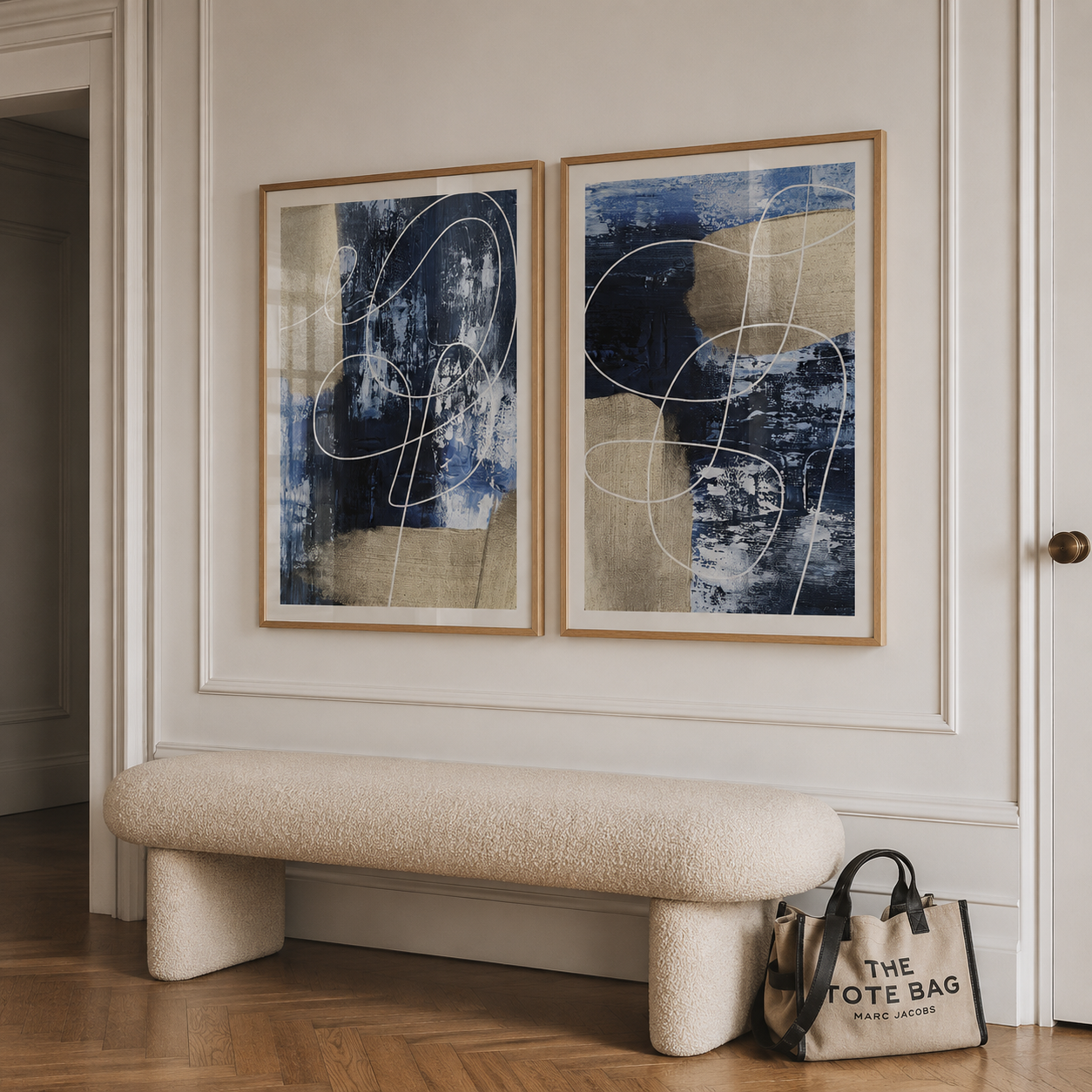 Two framed abstract canvases with blue, beige, and white tones hang on a cream wall above a textured cream boucle bench. A tote bag from The Tote Bag rests on the floor near the bench.