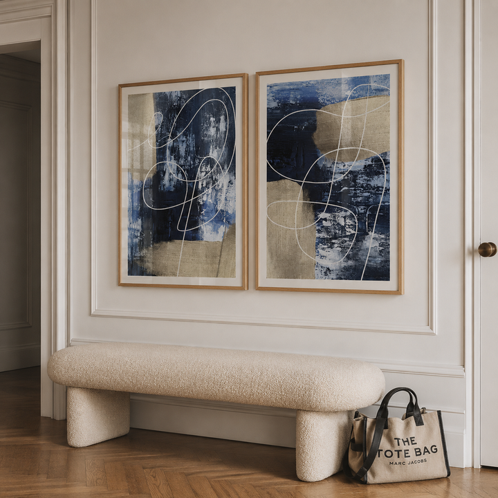 Two framed abstract canvases with blue, beige, and white tones hang on a cream wall above a textured cream boucle bench. A tote bag from The Tote Bag rests on the floor near the bench.