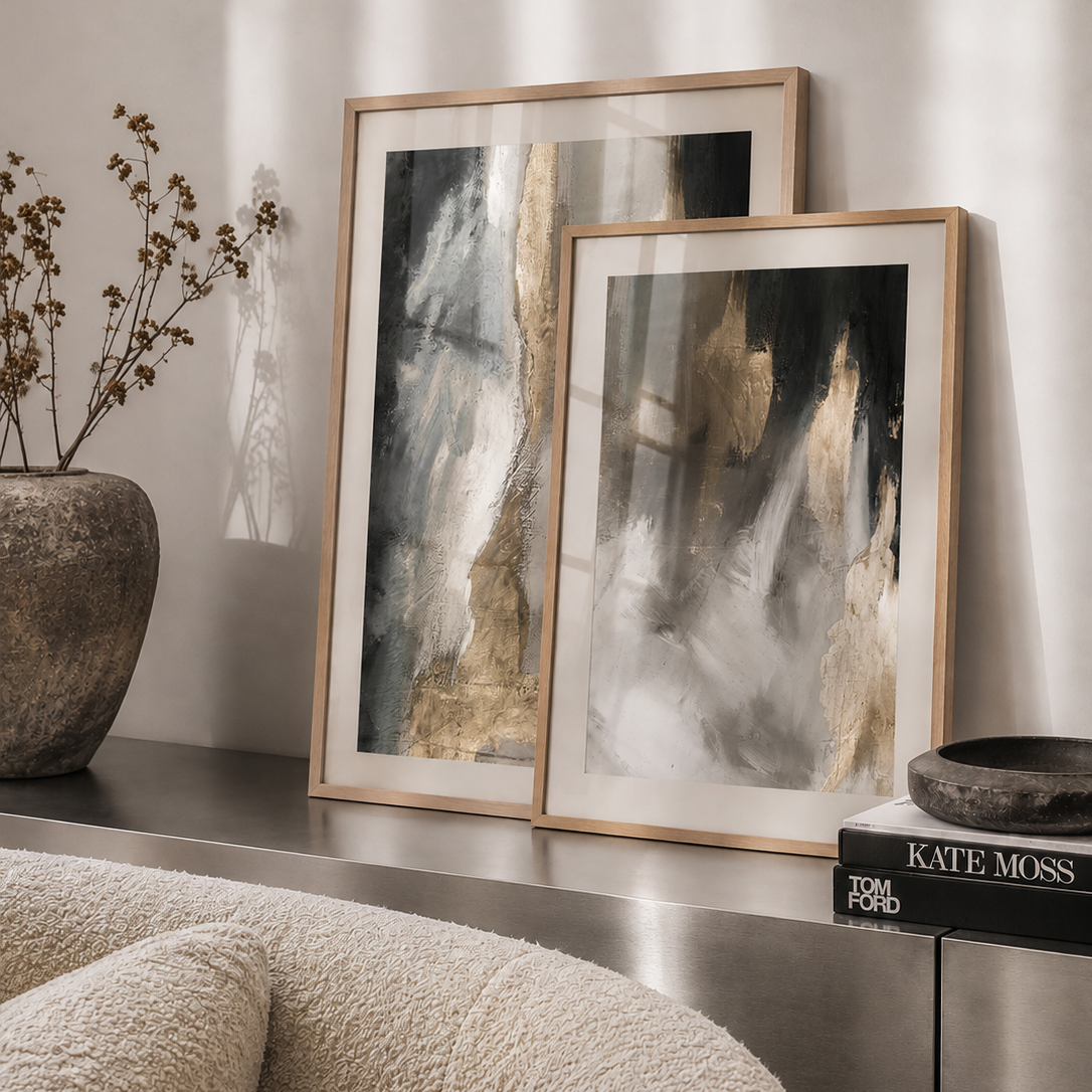 Two framed abstract wall art prints with gold and black brushstrokes, styled on a metallic console table next to a textured beige boucle chair and a stone vase.