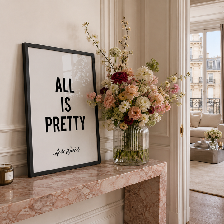 A framed black-and-white typographic print reading 'ALL IS PRETTY' with a small signature at the bottom, leaning on a pink marble console table beside a large floral arrangement in a ribbed glass vase. The room has cream walls, molding, and an open doorway leading to a sunlit living area.