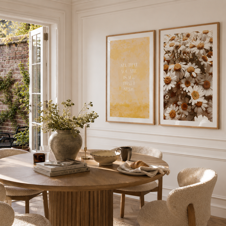 Two framed art prints hanging on a white paneled wall in a bright dining room. The left print has a textured mustard yellow background with elegant gold typography reading 'ALL THAT YOU ARE IS ALL THAT I NEED'.