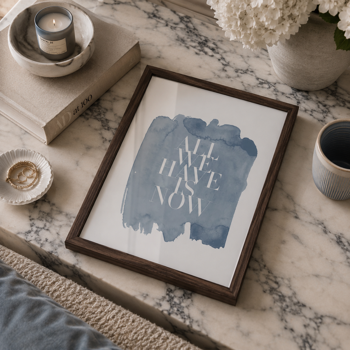 A framed art print featuring the text 'ALL WE HAVE IS NOW' in white over a blue watercolor splash, styled on a marble surface with a candle, books, flowers, and a cup nearby.