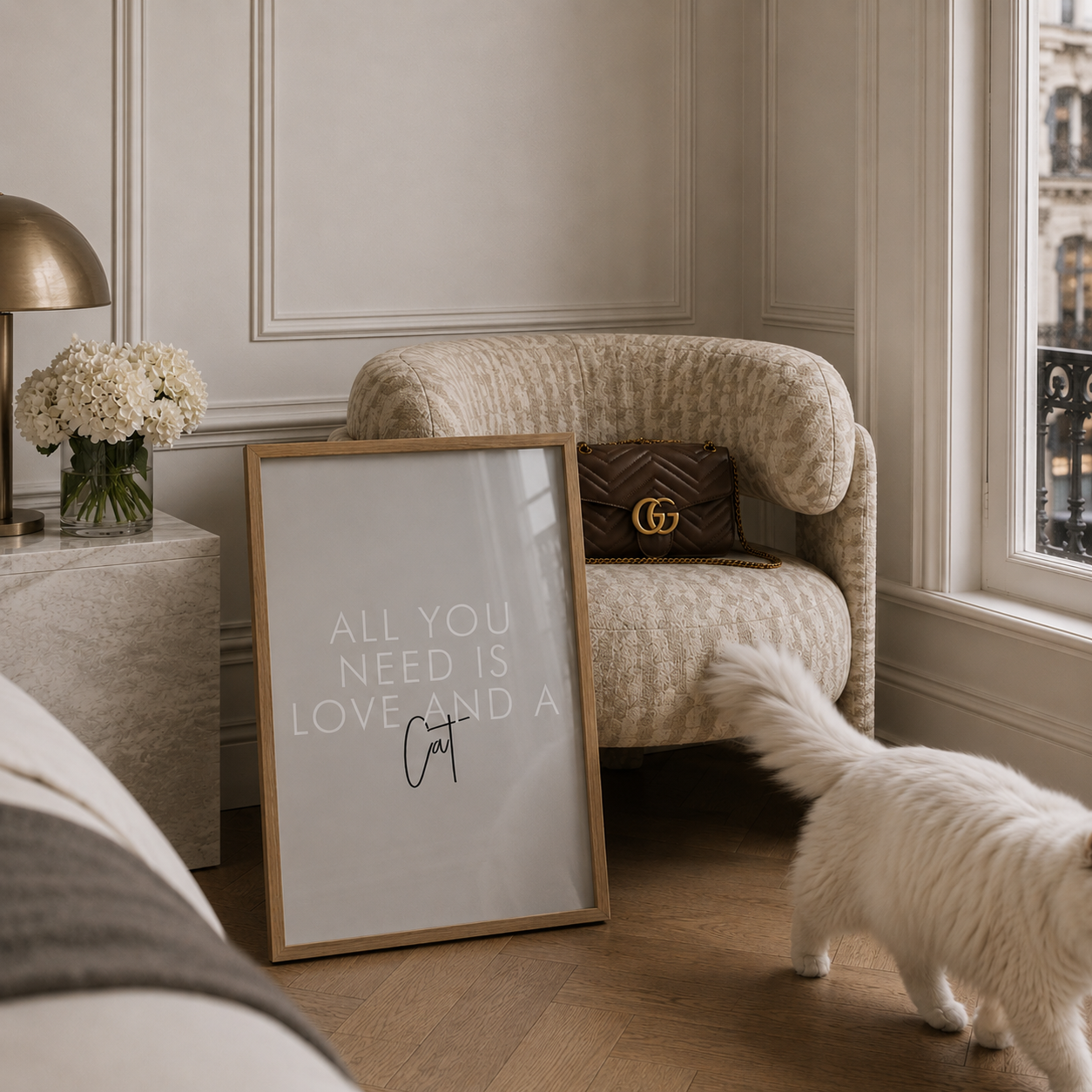 A framed art print with the text 'ALL YOU NEED IS LOVE AND A Cat' in white lettering on a light gray background, styled in a luxury room next to a beige armchair with a Gucci pillow and a white cat.