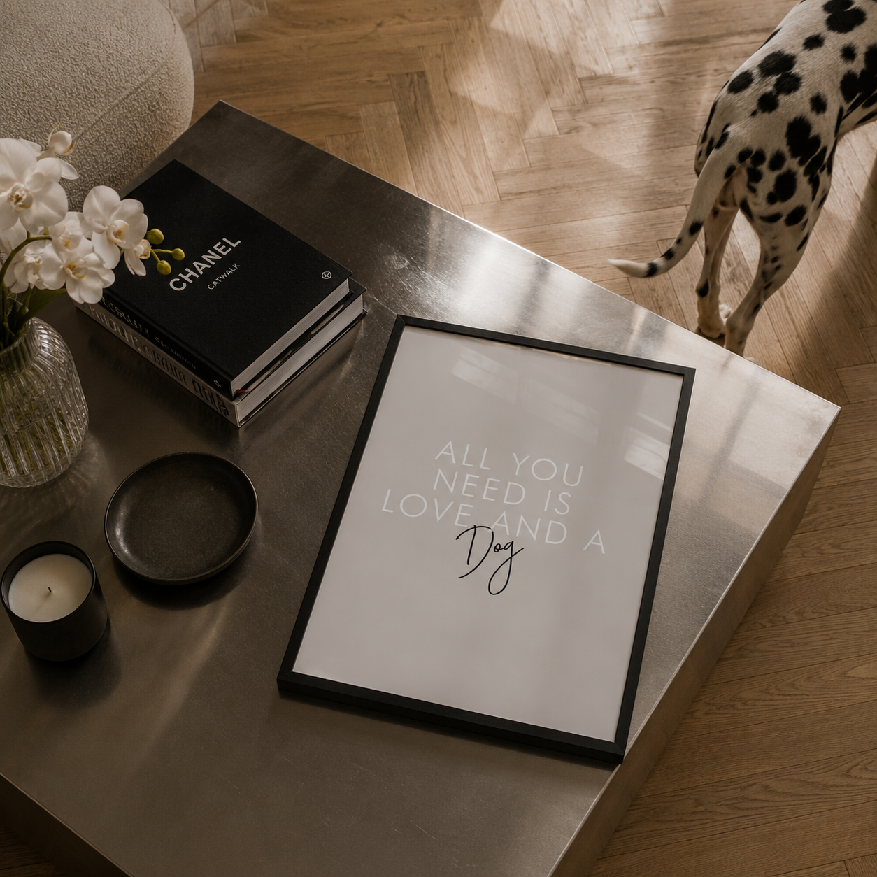 A framed minimalist art print with the text 'ALL YOU NEED IS LOVE AND A Dog' in white lettering on a light background, styled on a metallic coffee table next to a Chanel book, a candle, and a small dish, with a Dalmatian dog's tail visible on a wooden floor.