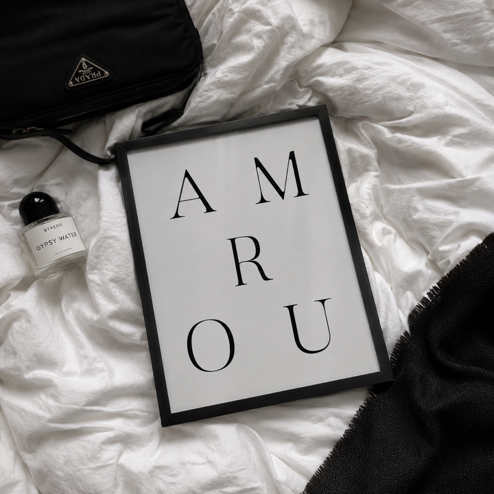 A framed typographic wall art print reading 'AMROU' in black serif letters on a white background, styled on rumpled white bedding with a black bag, a small perfume bottle, and a black textile nearby.