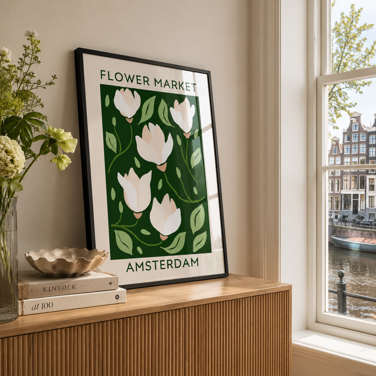 A framed art print featuring a stylized illustration of white tulips on a dark green background with the text 'FLOWER MARKET AMSTERDAM', leaning on a wooden console table next to a window with a canal view.