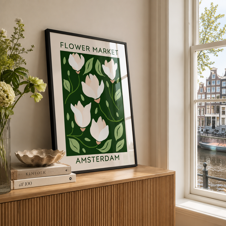 A framed art print featuring a stylized illustration of white tulips on a dark green background with the text 'FLOWER MARKET AMSTERDAM', leaning on a wooden console table next to a window with a canal view.