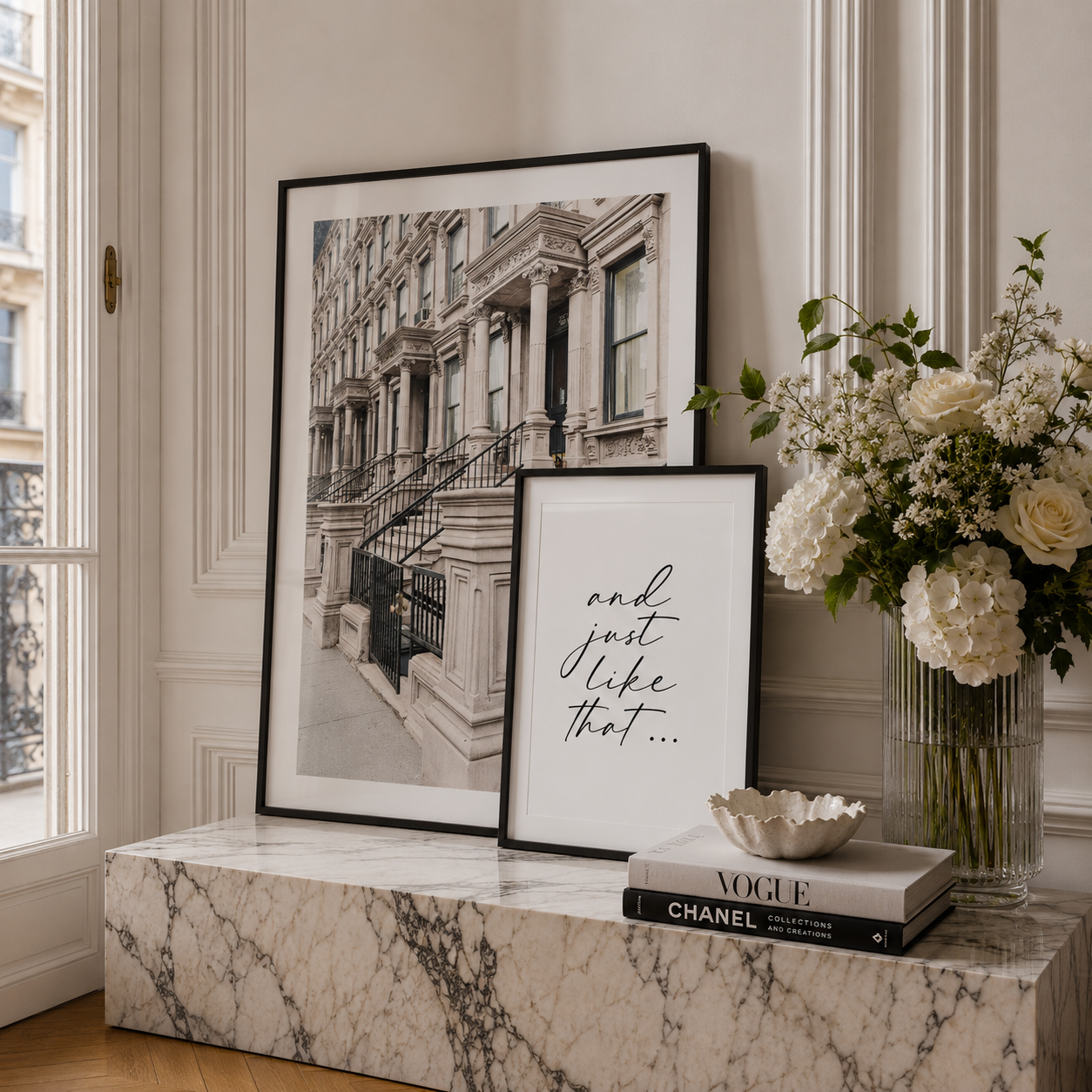 Two framed 'Sex and the City' inspired wall art prints displayed on a marble console in a bright, elegant room. One print is a black-and-white photograph of a classic brownstone building, and the other features elegant black script text on a white background.