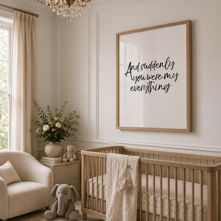A framed nursery wall print with the phrase 'And suddenly you were my everything' in black script inside a light wood frame, hanging above a wooden crib in a softly styled nursery with cream furniture, curtains, and a plush elephant.