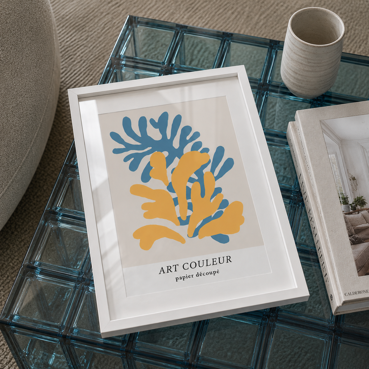 A framed abstract art print with blue and yellow organic shapes on a light beige background, titled 'ART COULEUR papier decoupe', displayed on a glass block coffee table next to a ceramic cup and a book.