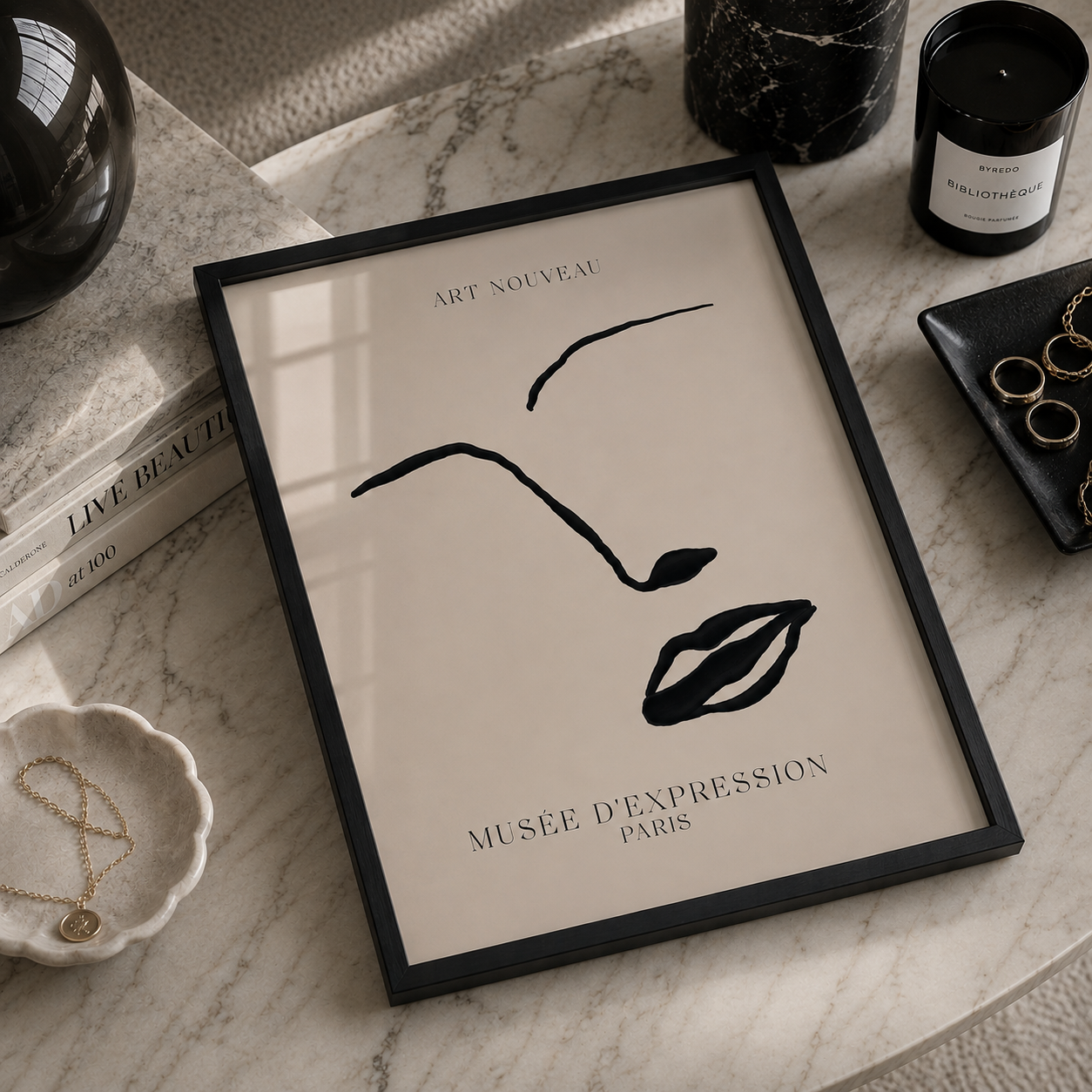 A framed minimalist art print featuring a black line drawing of a face and lips on a warm cream background, styled on a marble surface with books, candles, jewelry, and a small dish around it.