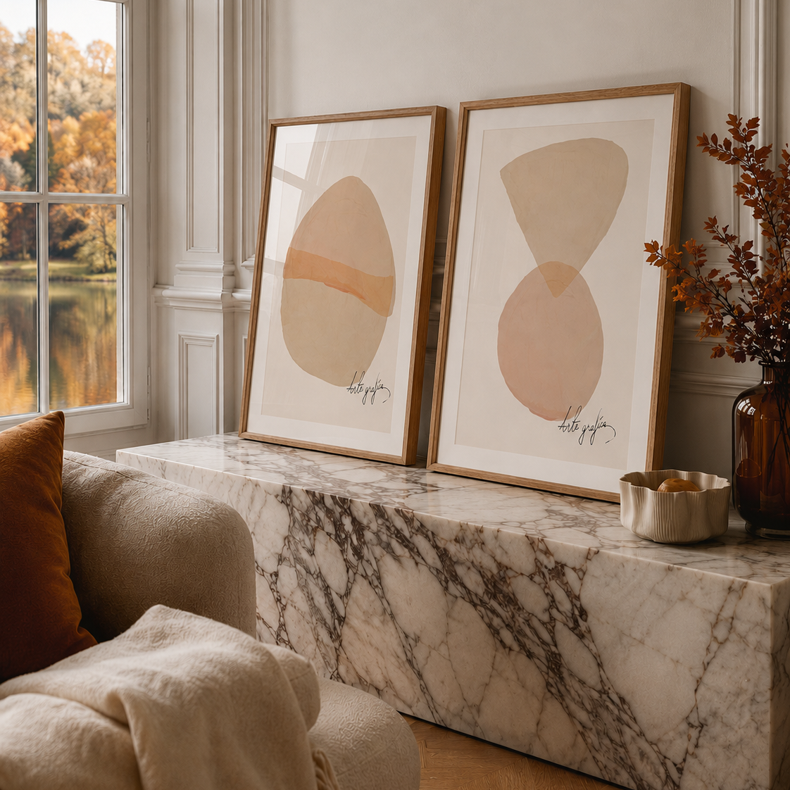 Two framed abstract art prints with soft beige and peach shapes in light wood frames, leaning on a marble console in a styled living room next to a window.