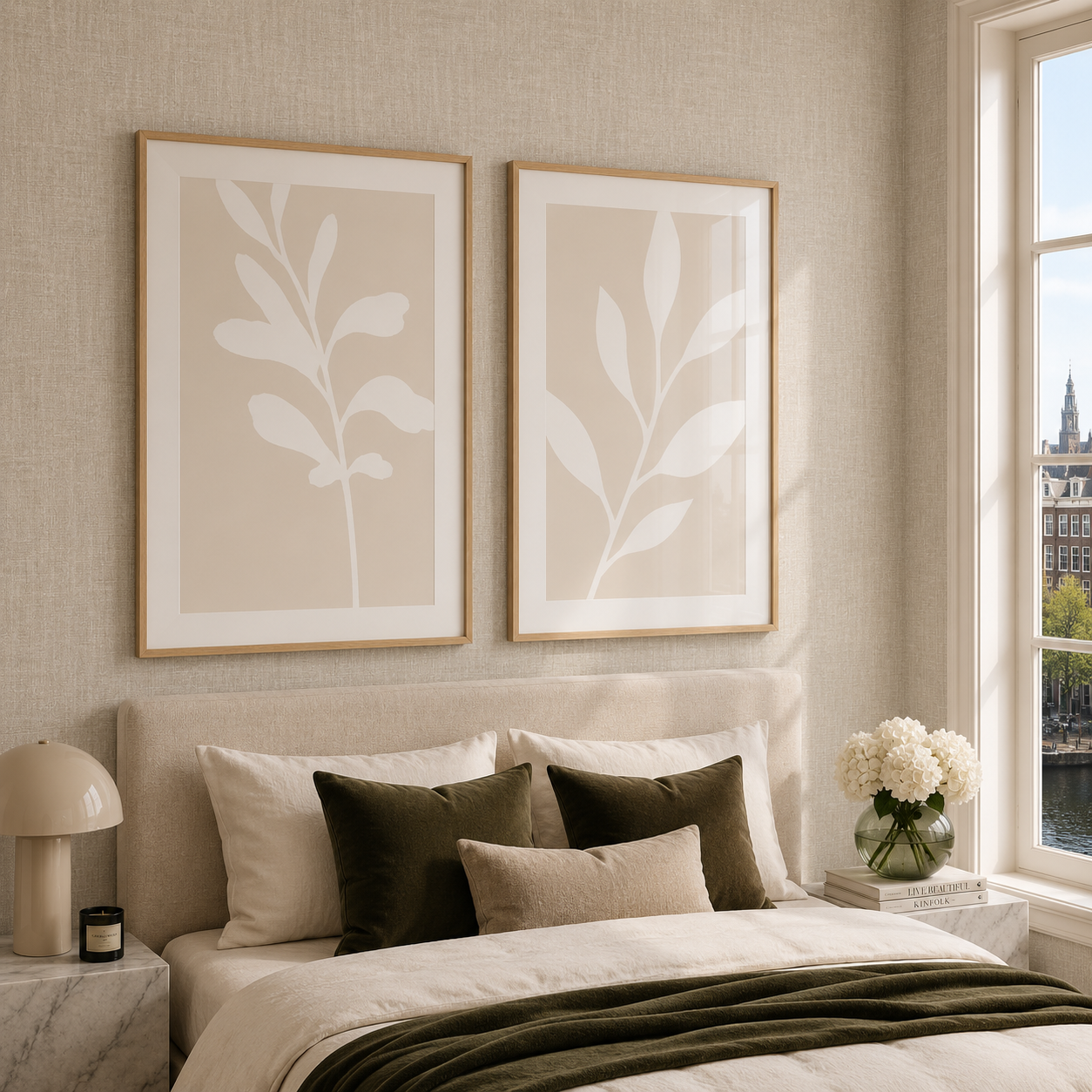A styled bedroom interior with two framed botanical wall art prints hanging above a bed. The prints feature large white leafy motifs on warm beige backgrounds with light wood frames.