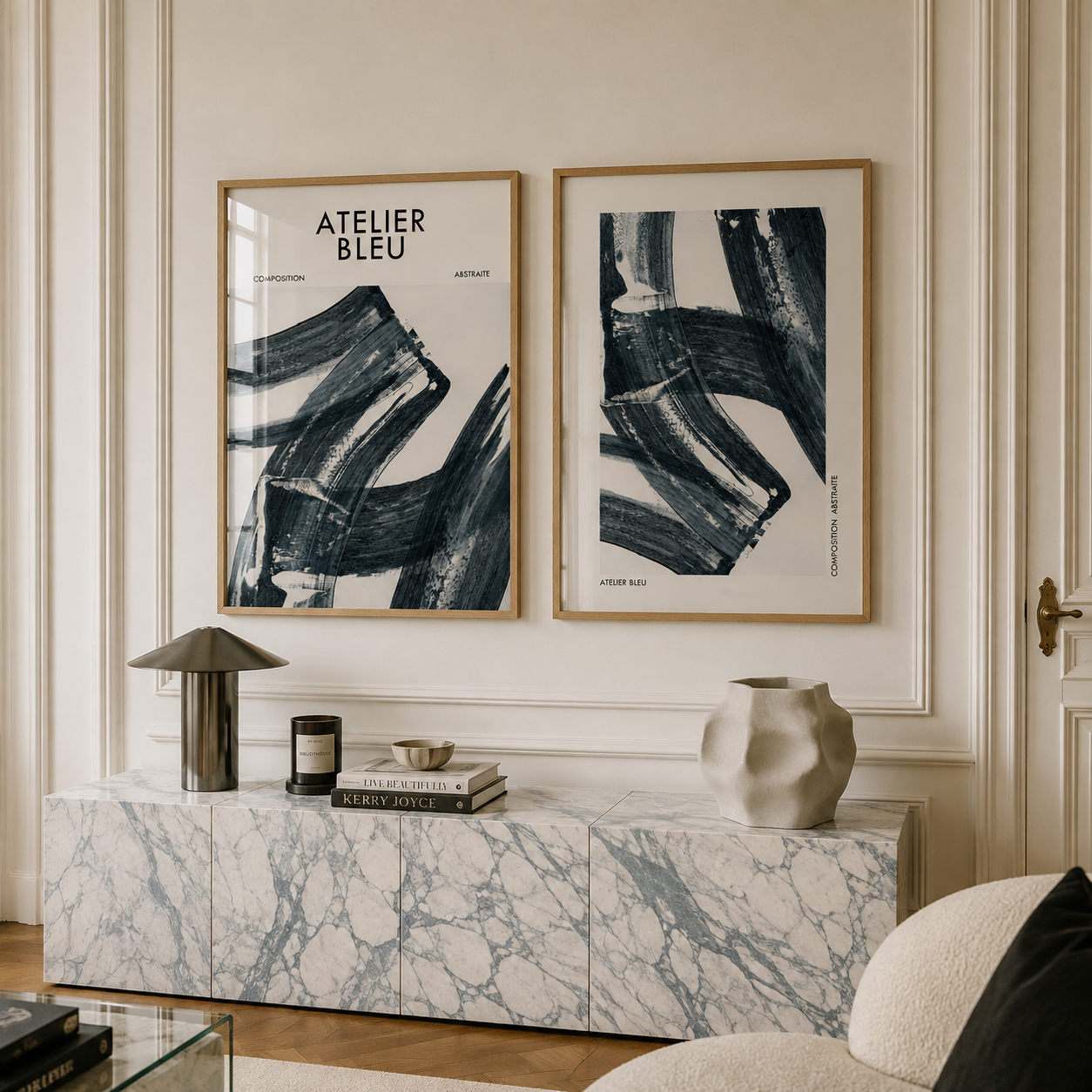 Two framed abstract art prints with black brushstrokes on a white background hang on a cream paneled wall above a marble console table. The left print reads 'ATELIER BLEU'.