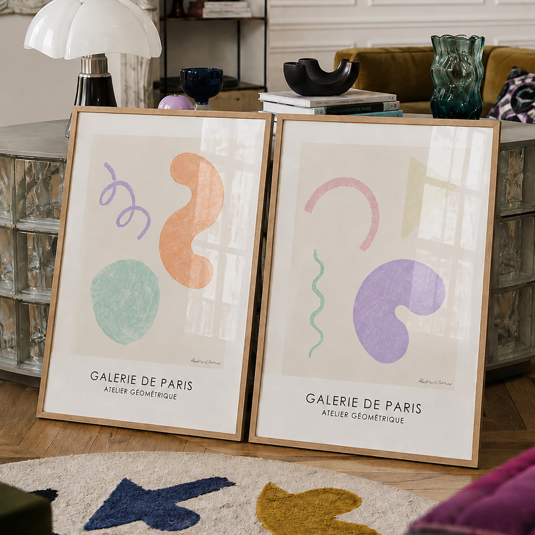 Two framed abstract art prints leaning against a mirrored sideboard in a styled living room. The prints feature pastel organic shapes and the text 'GALERIE DE PARIS' and 'ARTIER GÉOMÉTRIQUE'.
