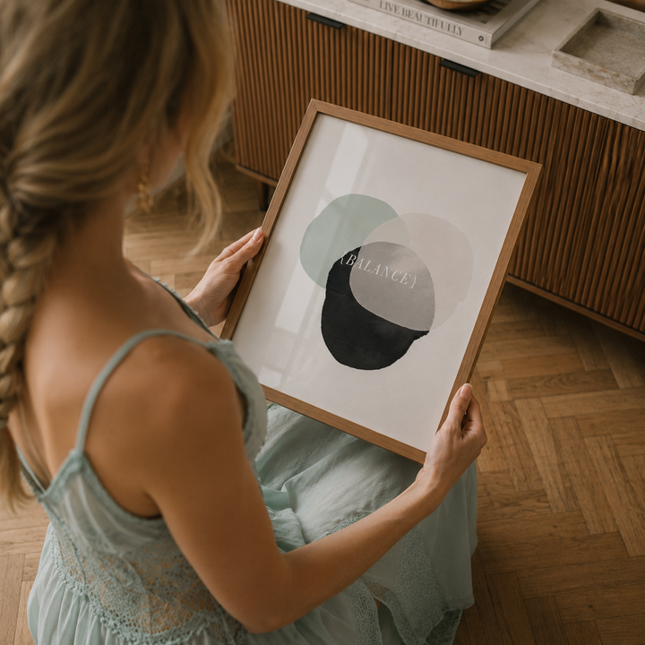 A woman in a light blue dress holds a framed abstract art print featuring overlapping circles in sage green, gray, and black with the word 'BELIEVE' written in white. The scene is set in a warm, modern room with wooden floors and slatted wood cabinetry.
