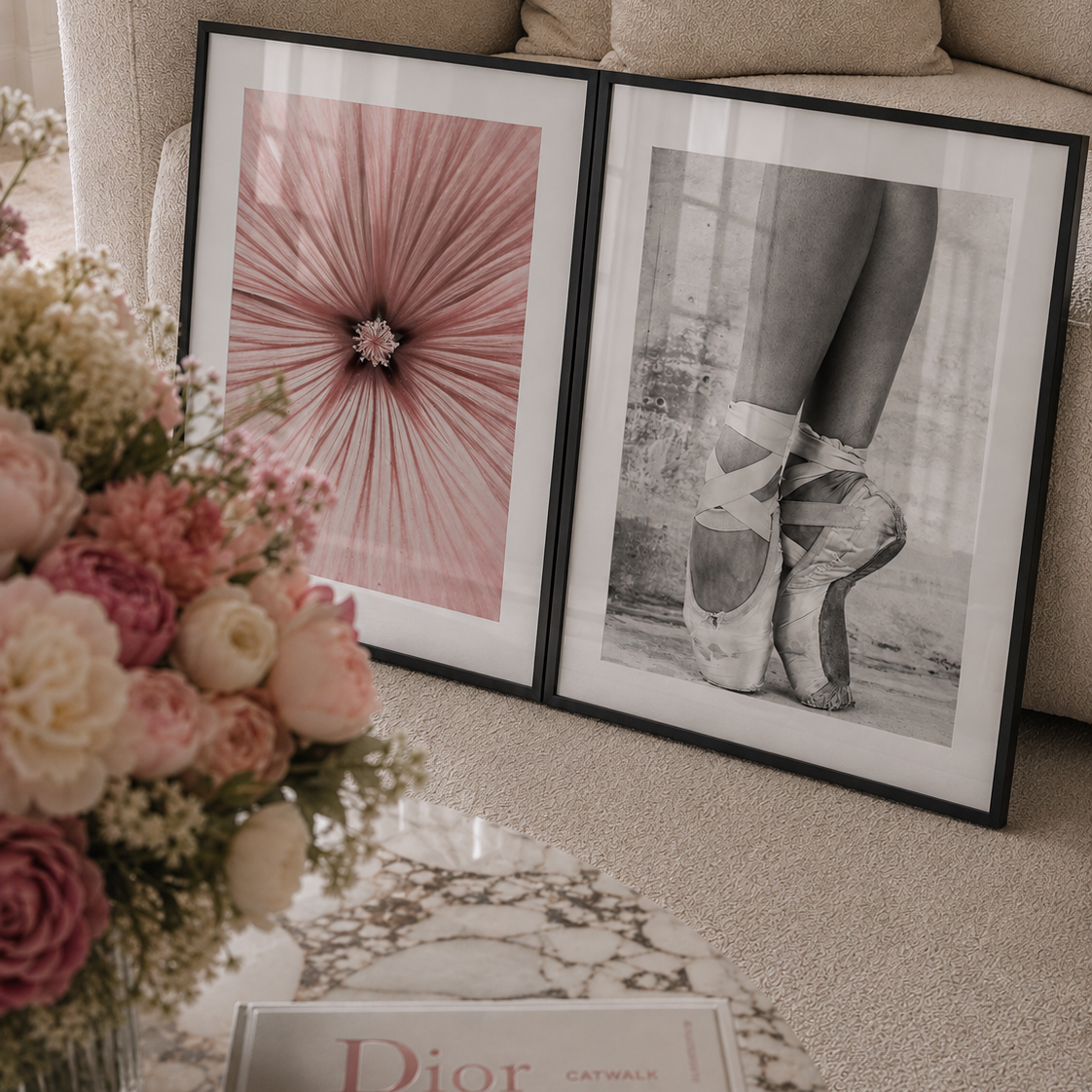 Two framed art prints leaning against a beige sofa in a styled living room. The left print shows a pink radial flower-like pattern, and the right print is a black-and-white photo of ballet pointe shoes.