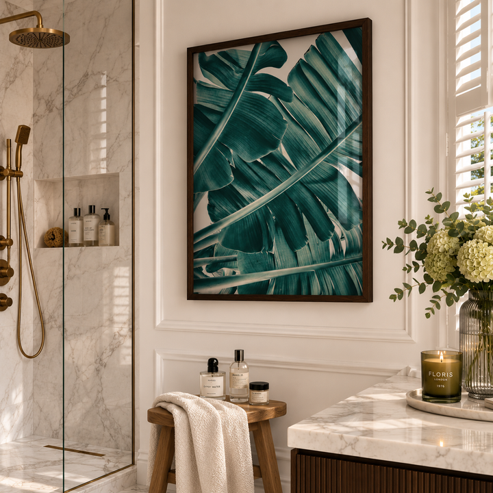 A framed wall art print featuring large teal-green tropical monstera leaves, displayed in a dark wood frame on a white paneled bathroom wall above a small wooden stool.