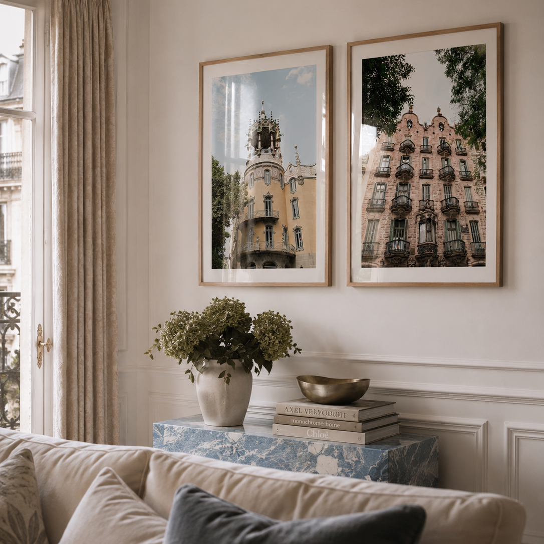 Two framed art prints depicting elegant European buildings hang on a cream-colored wall in a styled living room setting above a sofa with decorative cushions.