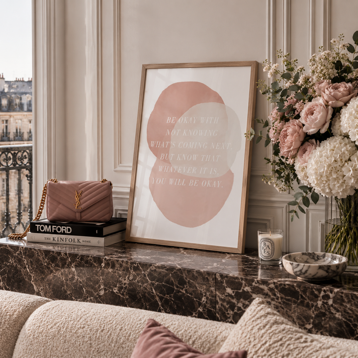 A framed art print with a minimalist pink and beige circle design and the text 'BE OK WITH NOT KNOWING WHAT'S COMING NEXT' displayed on a marble console table in a styled living room setting.