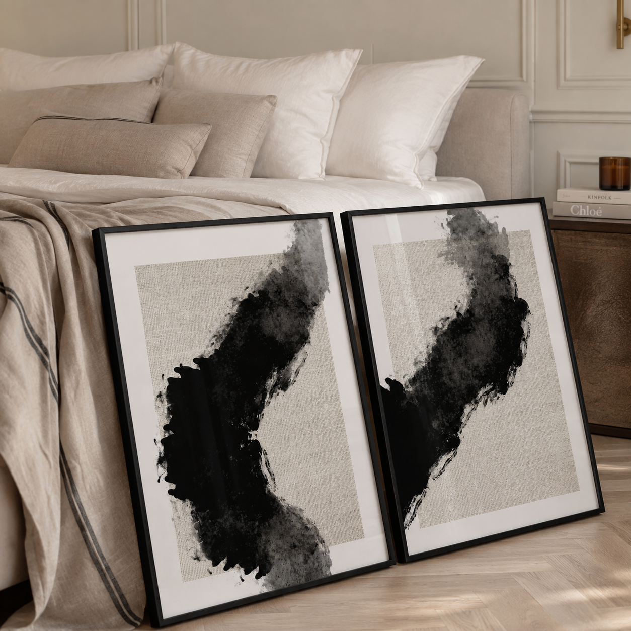 Two framed abstract black and gray watercolor art prints leaning against a bed in a styled bedroom setting.