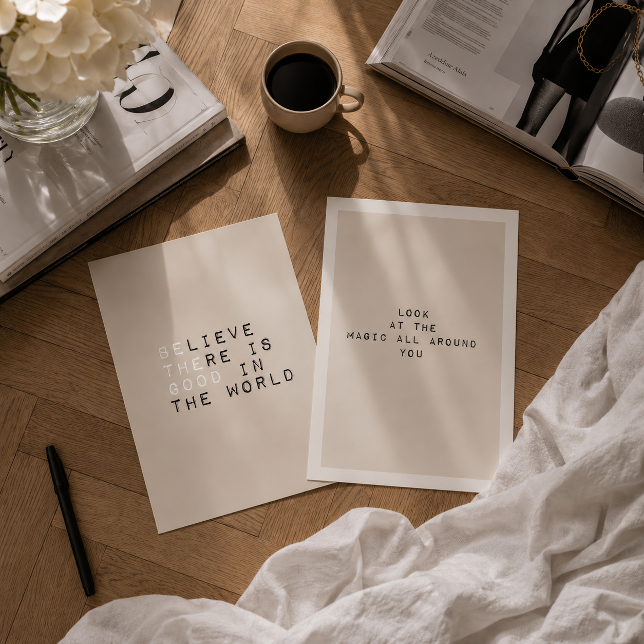 Two minimalist art prints with black text on a warm off-white background, styled on a wooden floor with a coffee cup, books, a pen, and white fabric.