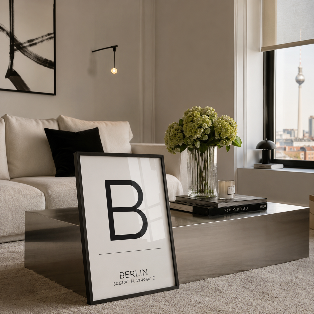 A framed minimalist art print featuring a large black letter 'B' and the text 'BERLIN' with coordinates, styled in a modern living room on a metallic coffee table next to a sofa and a vase of flowers.