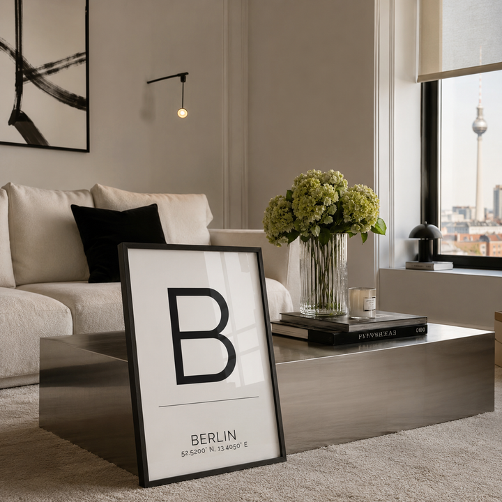 A framed minimalist art print featuring a large black letter 'B' and the text 'BERLIN' with coordinates, styled in a modern living room on a metallic coffee table next to a sofa and a vase of flowers.