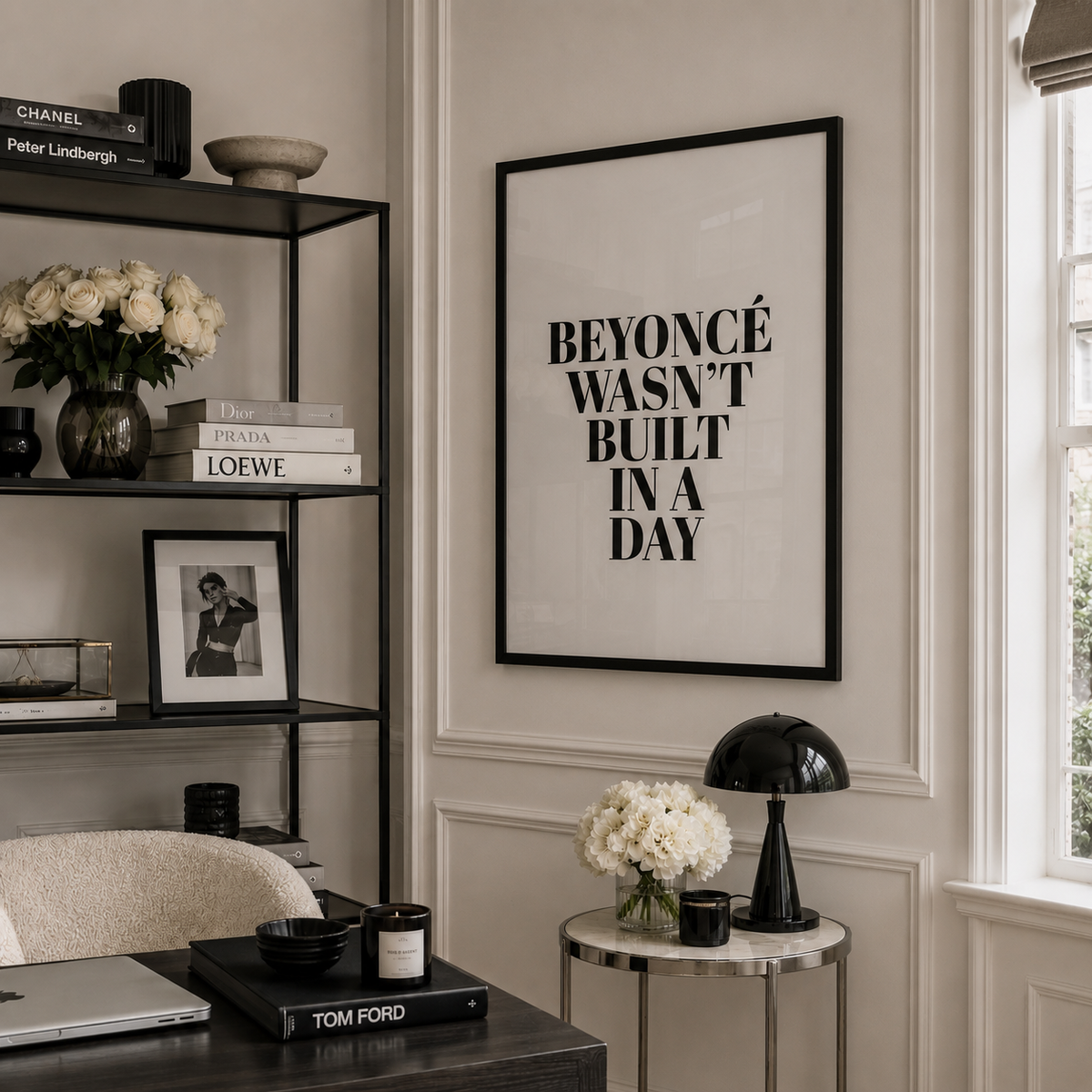 A framed black-and-white typographic wall print reading “BEYONCÉ WASN'T BUILT IN A DAY” hangs on a cream paneled wall in a styled home office. The room includes a black shelving unit with books and flowers, a desk, a side table with a black lamp and flowers, and a window.