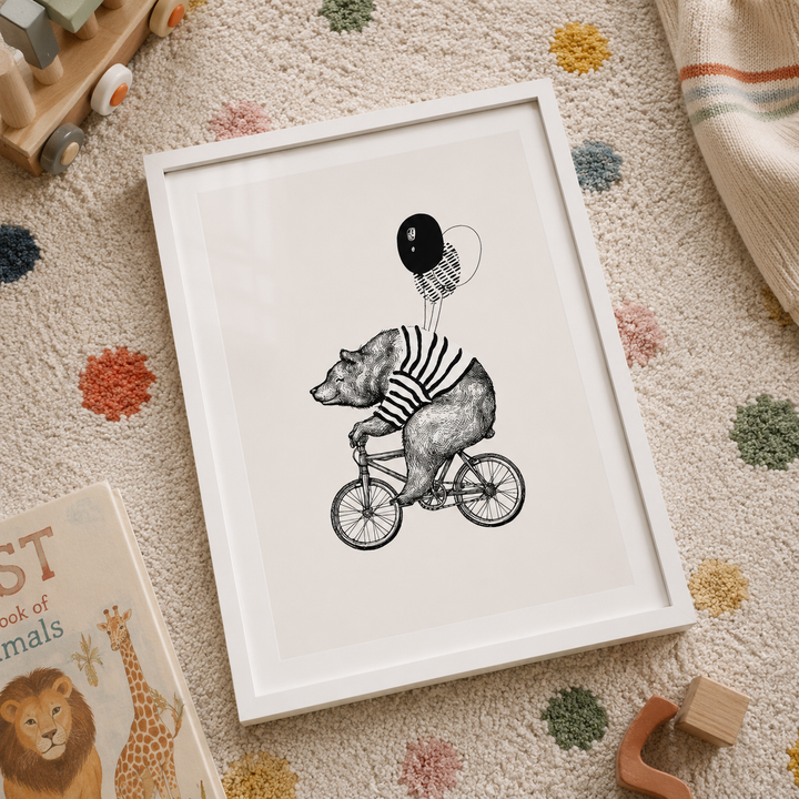 A framed art print featuring a black and white illustration of a bear riding a bicycle while holding balloons, displayed on a textured beige carpet with children's toys and a book nearby.