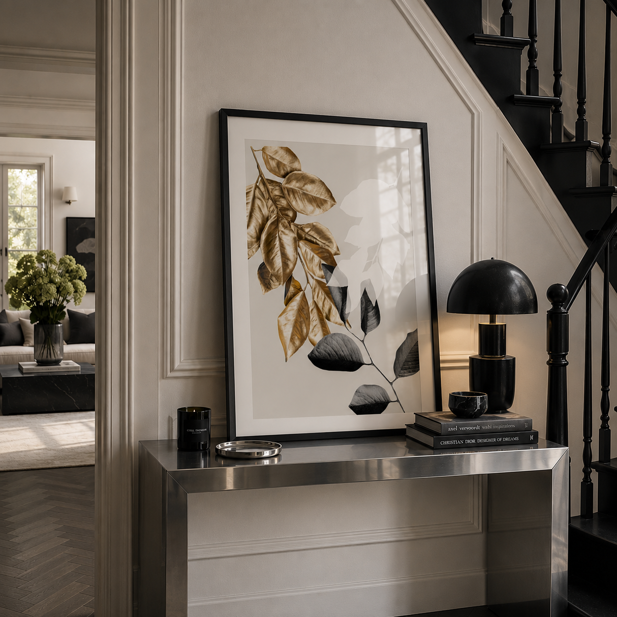 A framed botanical art print with gold and black leaves on a white background, styled on a silver console table in a modern home interior next to a black lamp and staircase.