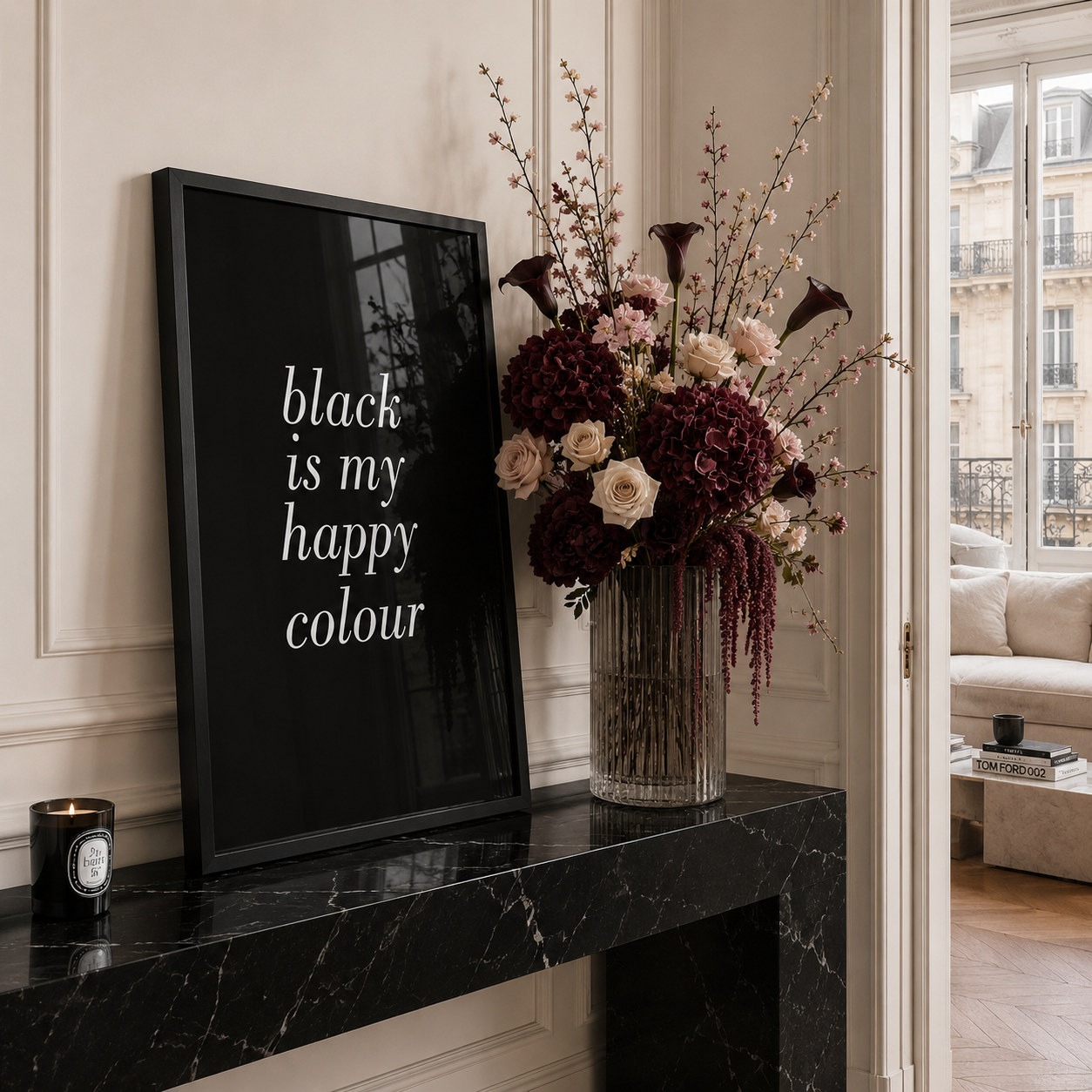 A framed black art print with white text reading 'black is my happy colour' sits on a black marble mantelpiece next to a large floral arrangement in a glass vase. The room has cream paneled walls and a window looking out onto a classic Parisian street.