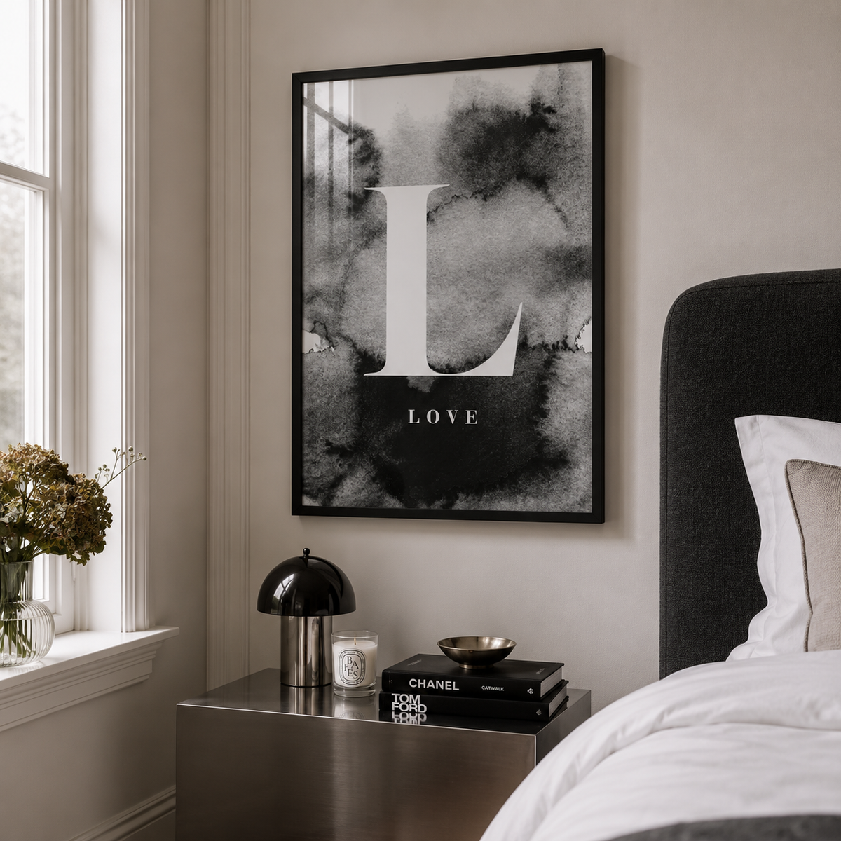 A framed black and white art print featuring a large letter 'L' and the word 'LOVE' in a bedroom setting, hanging on a beige wall above a metallic nightstand with a lamp and books.