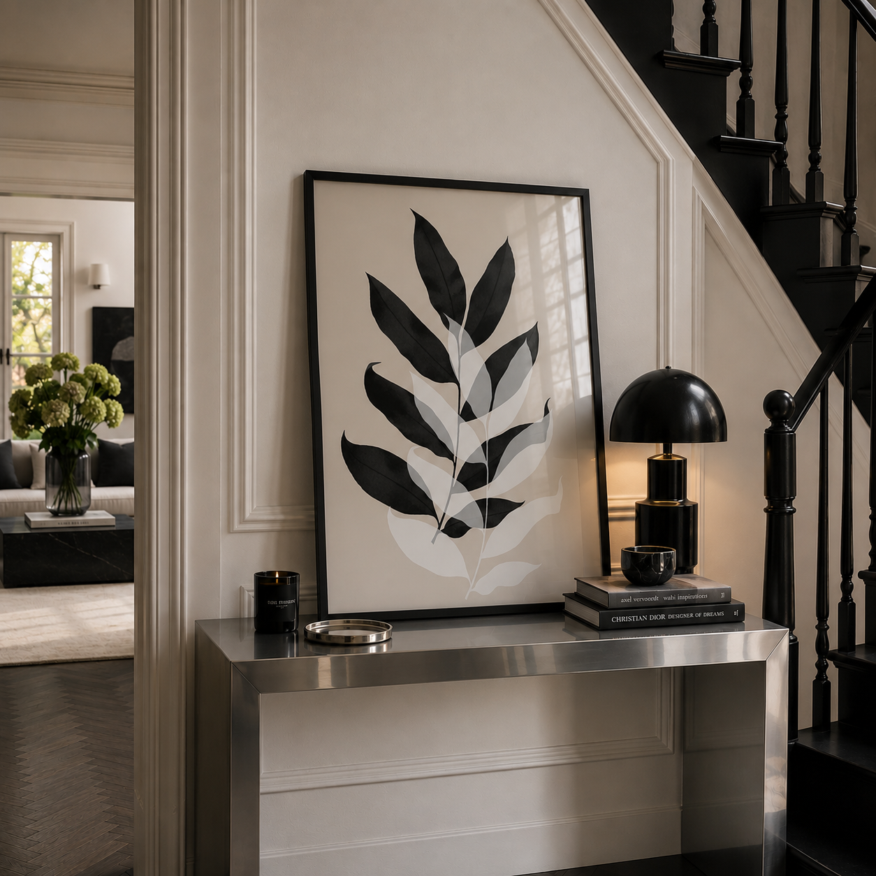 A large framed botanical art print with black and white leaf motifs hangs on a cream paneled wall above a metallic console table in a styled home interior next to a black staircase.