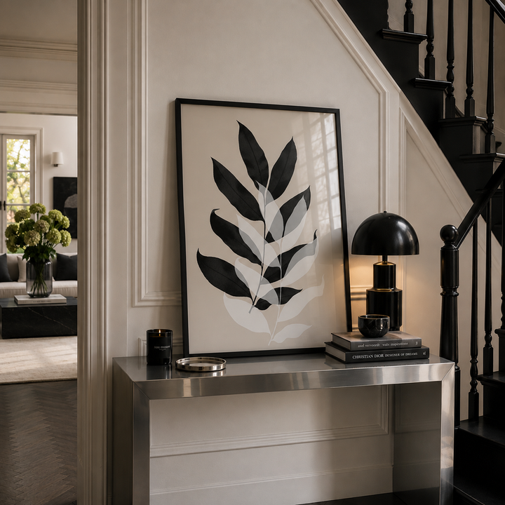 A large framed botanical art print with black and white leaf motifs hangs on a cream paneled wall above a metallic console table in a styled home interior next to a black staircase.