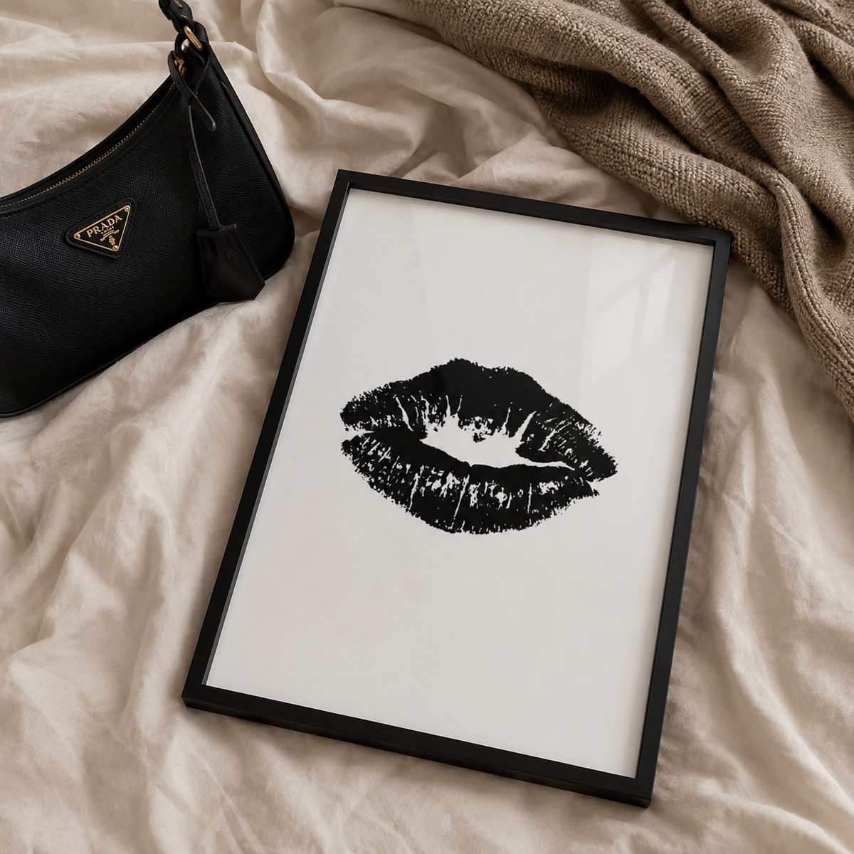 A framed black-and-white lip print art piece displayed on beige fabric, with a black handbag and a brown knit blanket nearby.