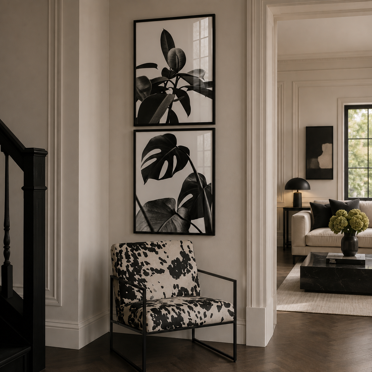 A styled interior entryway with two black-framed botanical wall prints above a black-and-white patterned armchair. A doorway leads to a living room with a sofa, lamp, and coffee table.