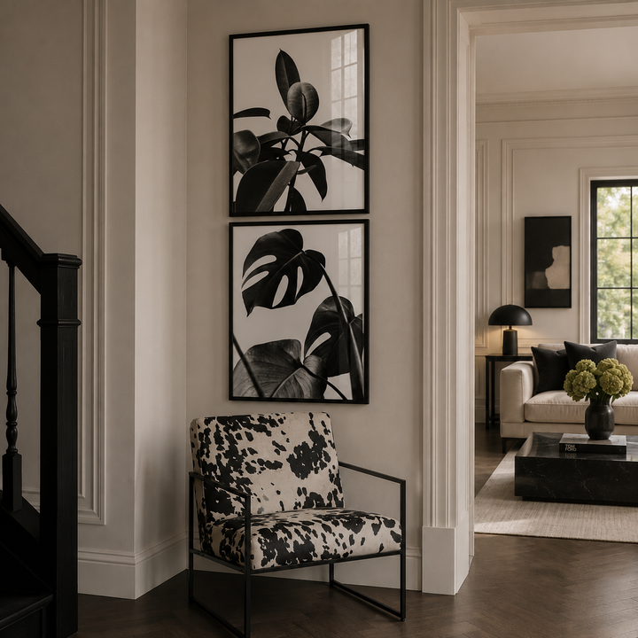 A styled interior entryway with two black-framed botanical wall prints above a black-and-white patterned armchair. A doorway leads to a living room with a sofa, lamp, and coffee table.