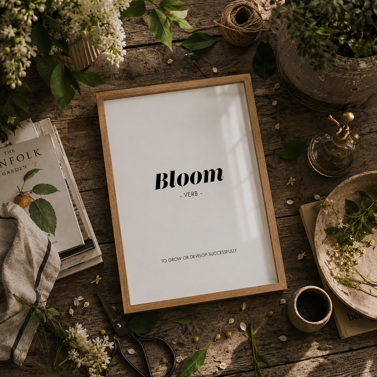 A framed typographic art print reading 'Bloom - VERB - TO GROW OR DEVELOP SUCCESSFULLY' lies on a rustic wooden table surrounded by flowers, scissors, a small cup, twine, and books.