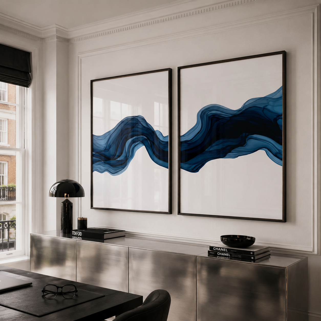 Two large framed abstract wall art prints with flowing blue and navy wave-like forms on a white background, displayed above a metallic console in a bright modern room.