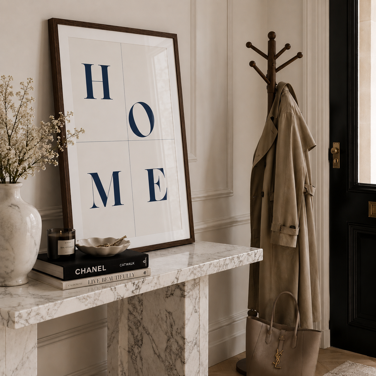 A framed wall art print with the word HOME spelled out in large navy letters on a white background, styled on a marble console table in a home entryway next to a coat rack with a trench coat and a handbag.