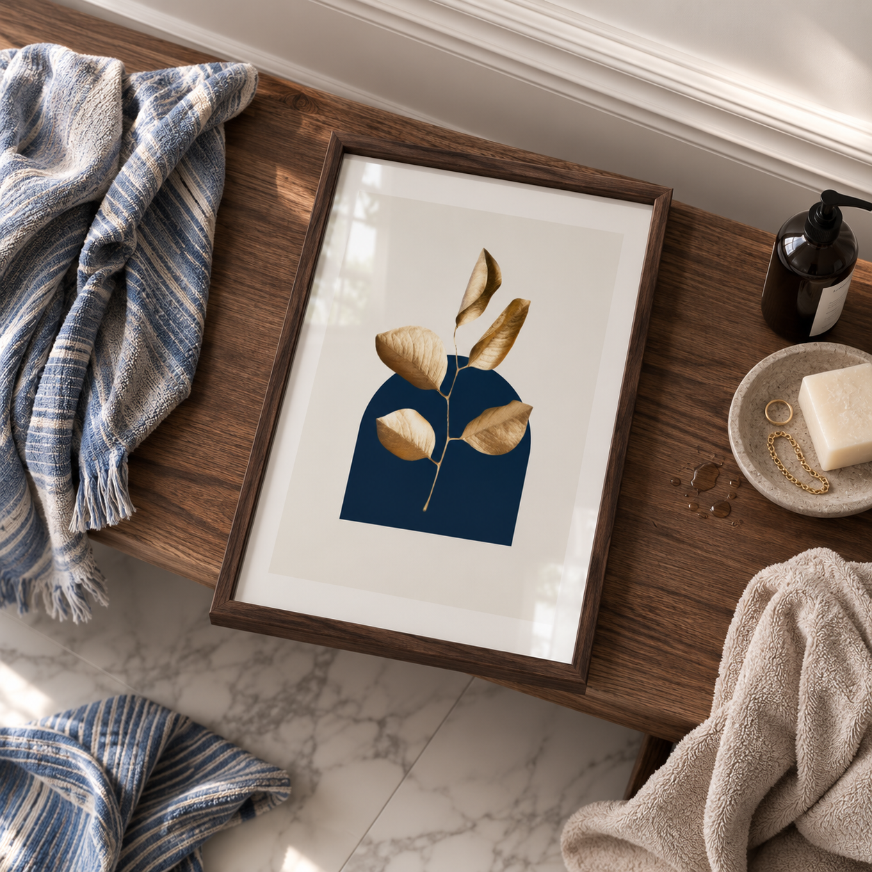 A framed botanical art print featuring gold leaves on a navy blue background, styled on a wooden bathroom bench with towels, soap, and accessories.